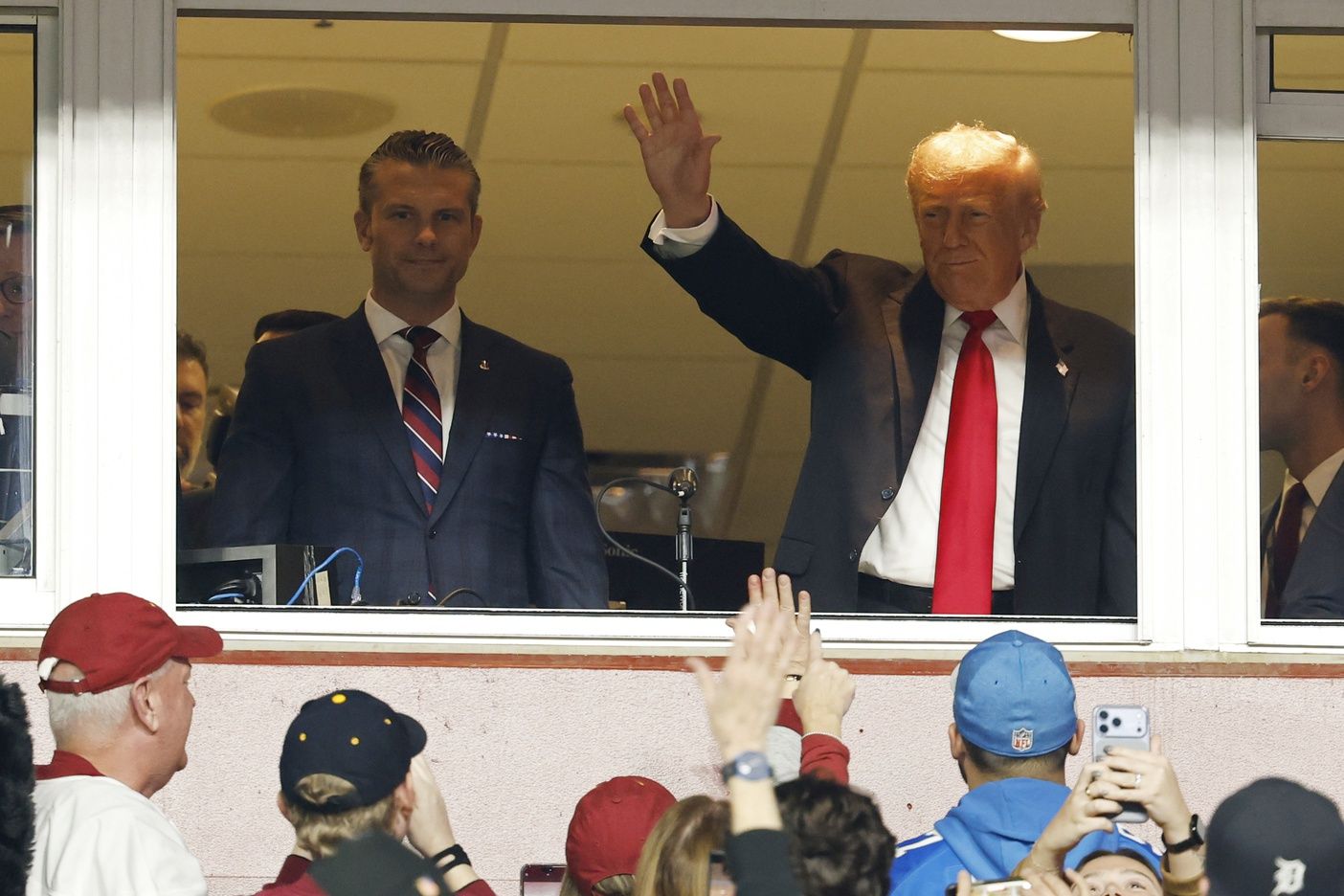 President Donald Trump waves to fans while standing next to U.S. Secretary of War Pete Hegseth (L) after swearing in a new group of U.S. military recruits during halftime of the game between the Detroit Lions and Washington Commanders as part of the NFL Salute to Service initiative at Northwest Stadium.