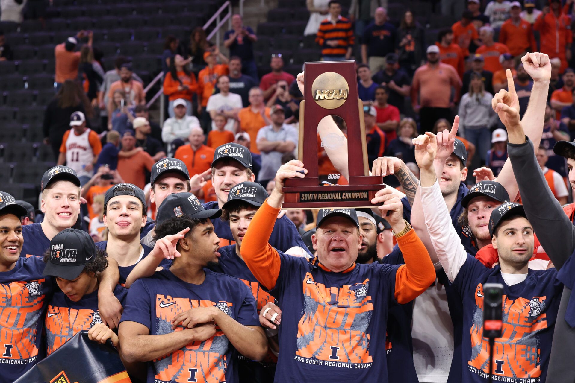 Illinois Fighting Illini head coach Brad Underwood and players celebrate with the trophy on the podium after defeating the Iowa Hawkeyes in an Elite Eight game of the South Regional of the men's 2026 NCAA Tournament at Toyota Center.